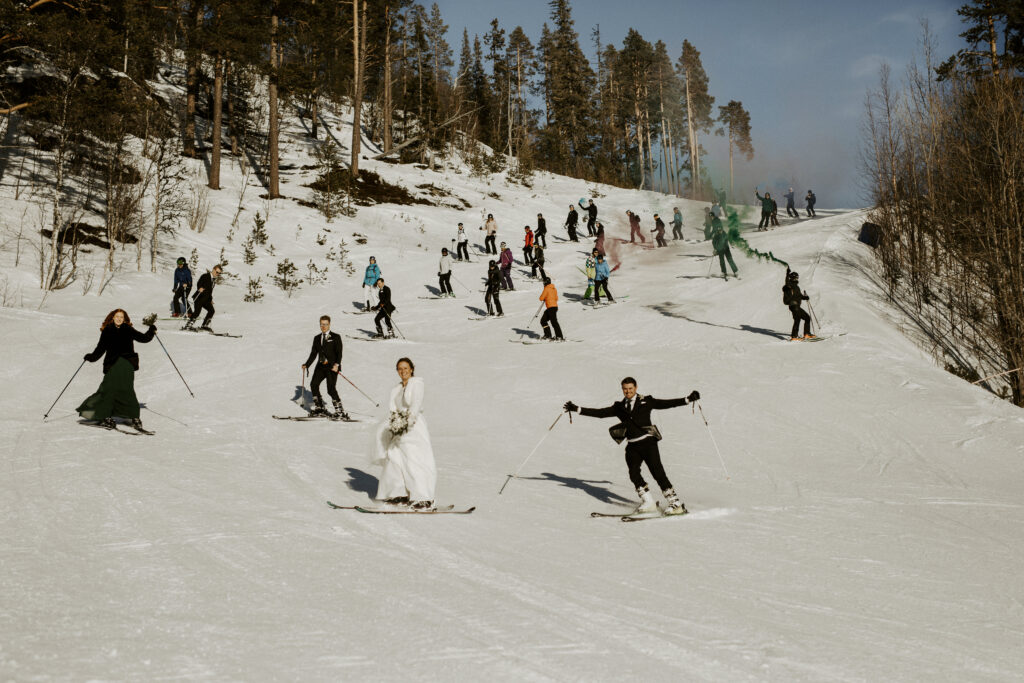 Bröllopsgäster och brudpar åker skidor nerför backe i fjällen